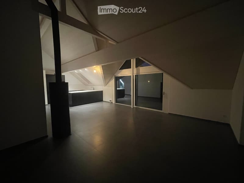 An empty attic space with a slanted ceiling, dark flooring, and large windows, illuminated by soft lighting.