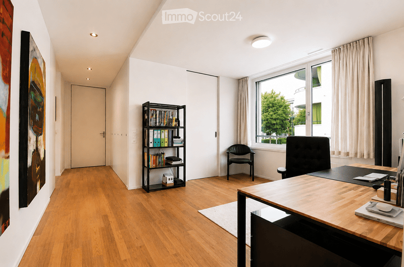 Modern office space with wooden flooring, large window, desk, chairs, bookshelf, and artwork on the wall.