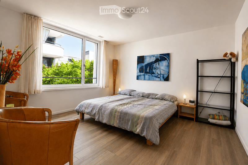 Modern bedroom with wooden flooring, large window with view, bed with patterned bedding, nightstand, chair, shelf, vase with flowers, and artwork on walls.
