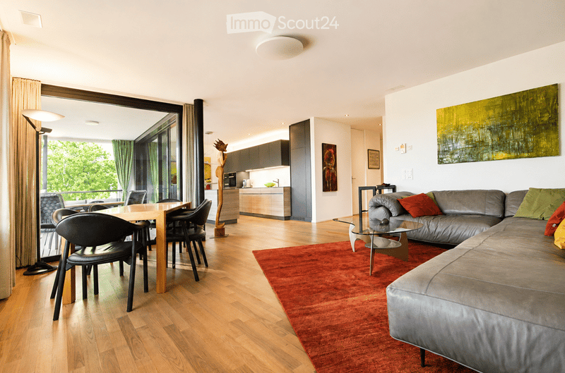 Modern open-plan living and dining area with hardwood flooring, large windows offering a view, a grey sectional sofa with colorful cushions, a red area rug, a glass coffee table, a wooden dining table with black chairs, and a sleek kitchen area in the bac