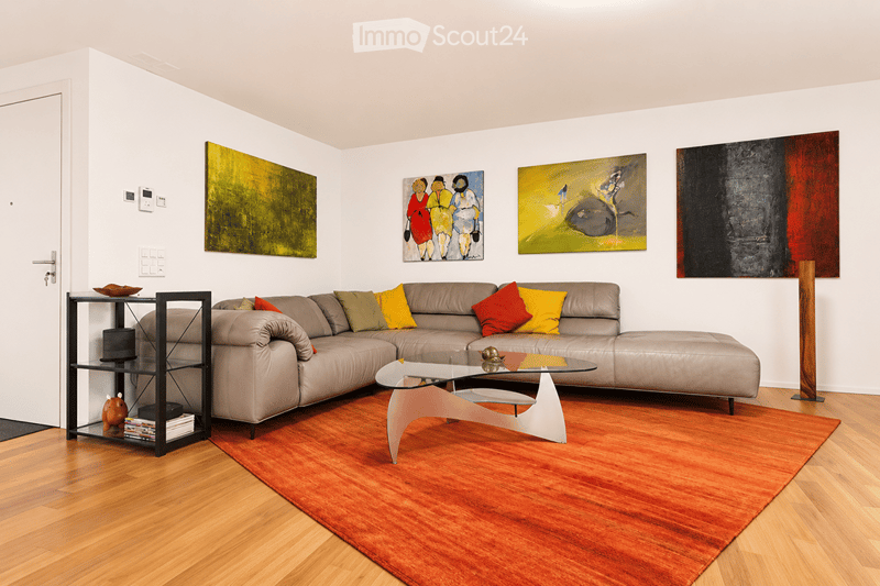 Modern living room with a grey sectional sofa, colorful throw pillows, a glass coffee table, a side table with books and a small figurine, and several artworks on the walls.