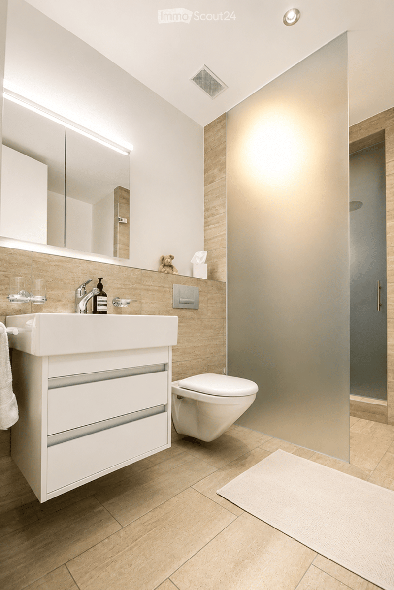 Modern bathroom with a white sink, mirror, toilet, and a glass shower stall. The floor is tiled, and there is a towel on the side.