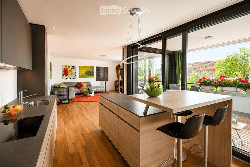 Modern open-plan kitchen and living area with wooden flooring, large windows offering a view, a kitchen island with bar stools, a sofa, coffee table, and decorative artwork.