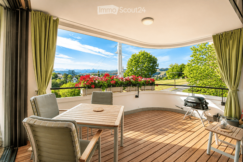 Wooden balcony with a table, chairs, and a grill, overlooking a scenic view with trees and a cityscape.