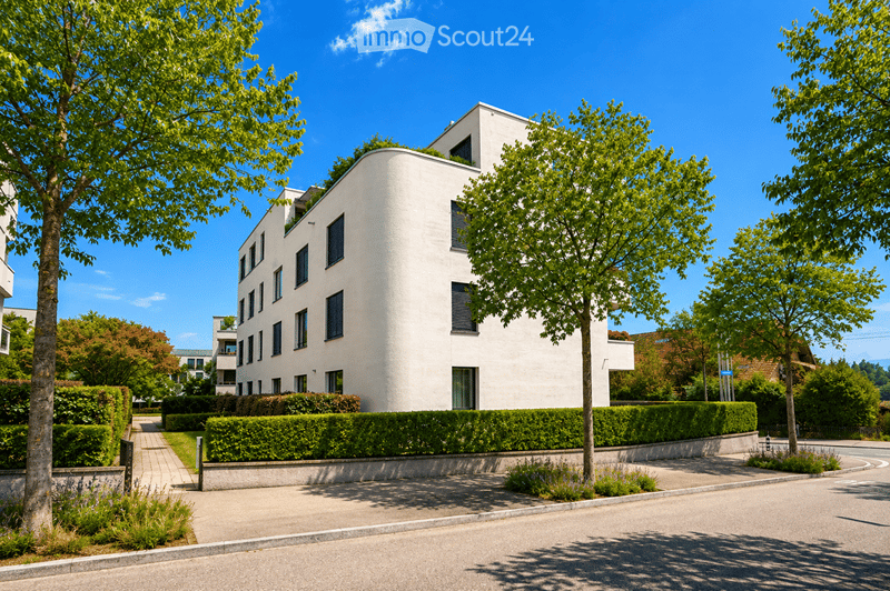 Modern white multi-story building with clean lines and numerous windows, surrounded by greenery and trees.
