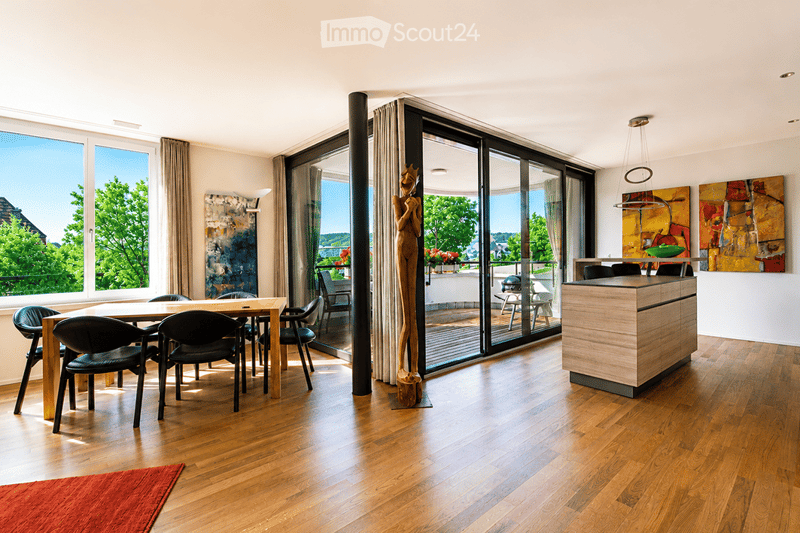 Bright, open-plan dining and kitchen area with wooden flooring, large windows offering a scenic view, a dining table with chairs, a kitchen island, and decorative art pieces.