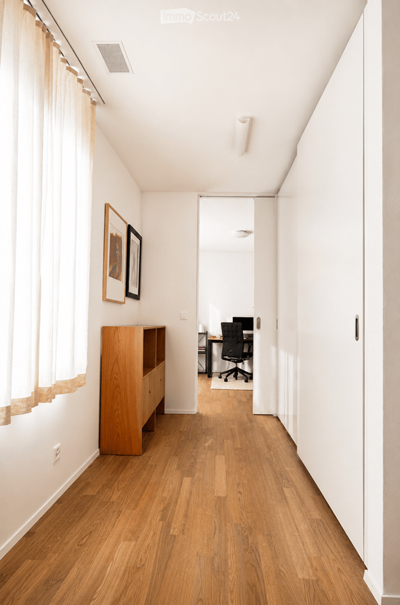 A narrow hallway with wooden flooring, white walls, and a wooden cabinet on the left side. A doorway leads to a room with a desk, chair, and computer.