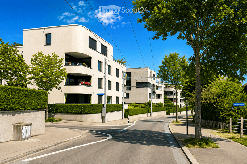 Modern multi-story residential building with balconies, surrounded by greenery and trees on a sunny day.