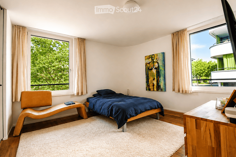 Modern bedroom with a double bed, wooden chair, desk, and large windows offering a view of greenery.