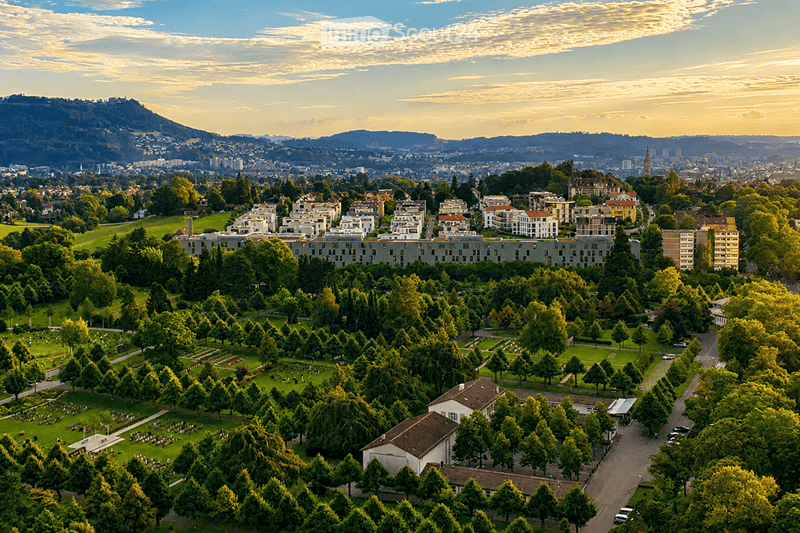 Aerial view of a cityscape with residential buildings, green spaces, and a distant mountain range under a clear sky.