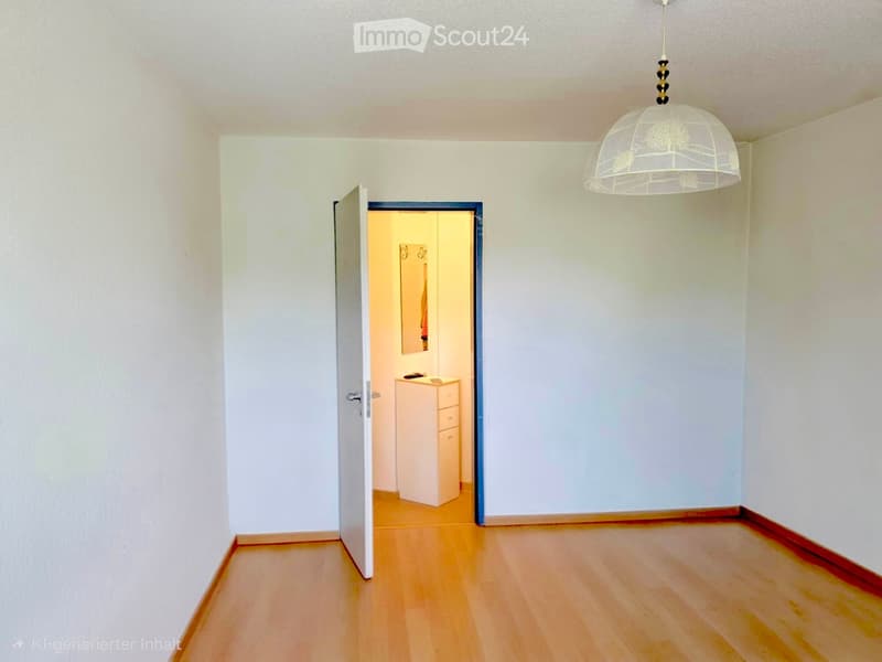 Empty hallway with light wooden flooring, white walls, and a hanging glass pendant light. An open door leads to another room with a small cabinet and mirror.