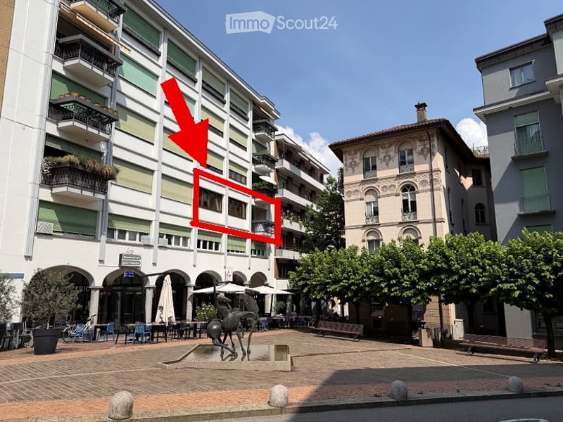 Multi-story apartment building with balconies and green plants, located in a town square with outdoor seating, tables, and a sculpture in the foreground.