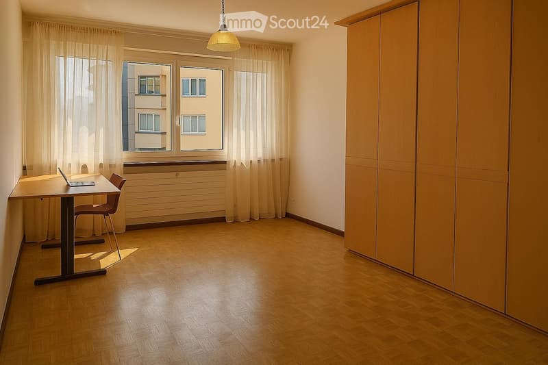 A furnished room with wooden flooring, a large window with sheer curtains, a table with a laptop, a chair, and a built-in wardrobe.