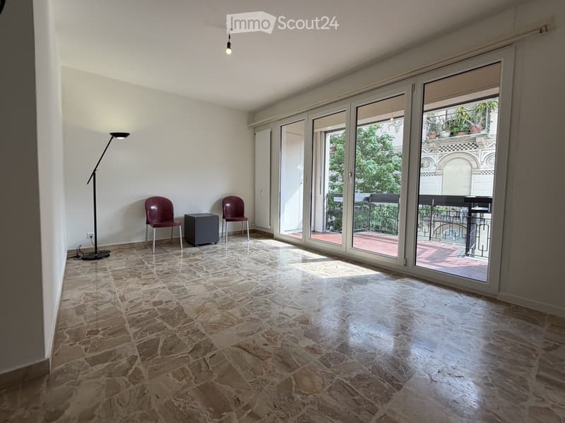 Bright, unfurnished room with large sliding glass doors leading to a balcony, stone tile flooring, and minimal furniture including two chairs, a small table, and a floor lamp.