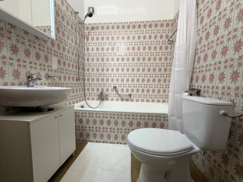 Bathroom with floral patterned tiles, a white sink, bathtub, toilet, and a small cabinet.