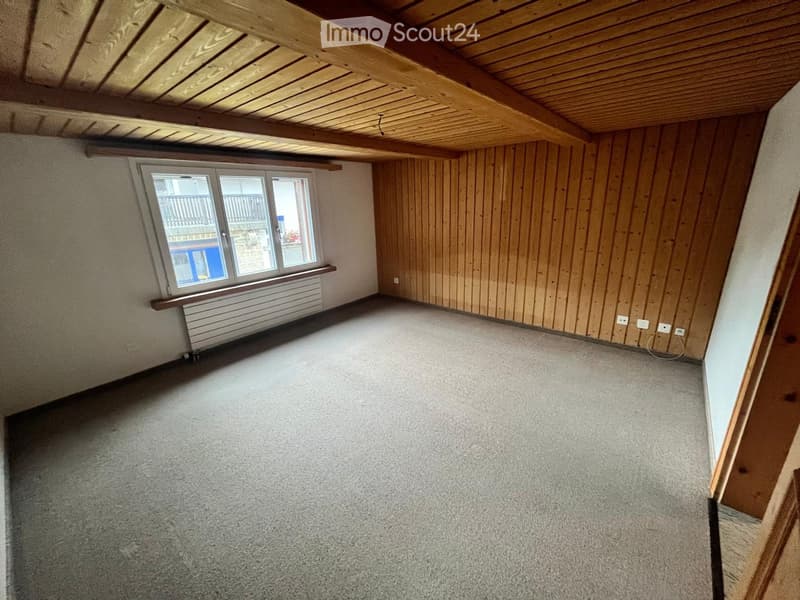 Empty attic room with wooden walls, exposed wooden beams, and a large window with a view of a balcony.