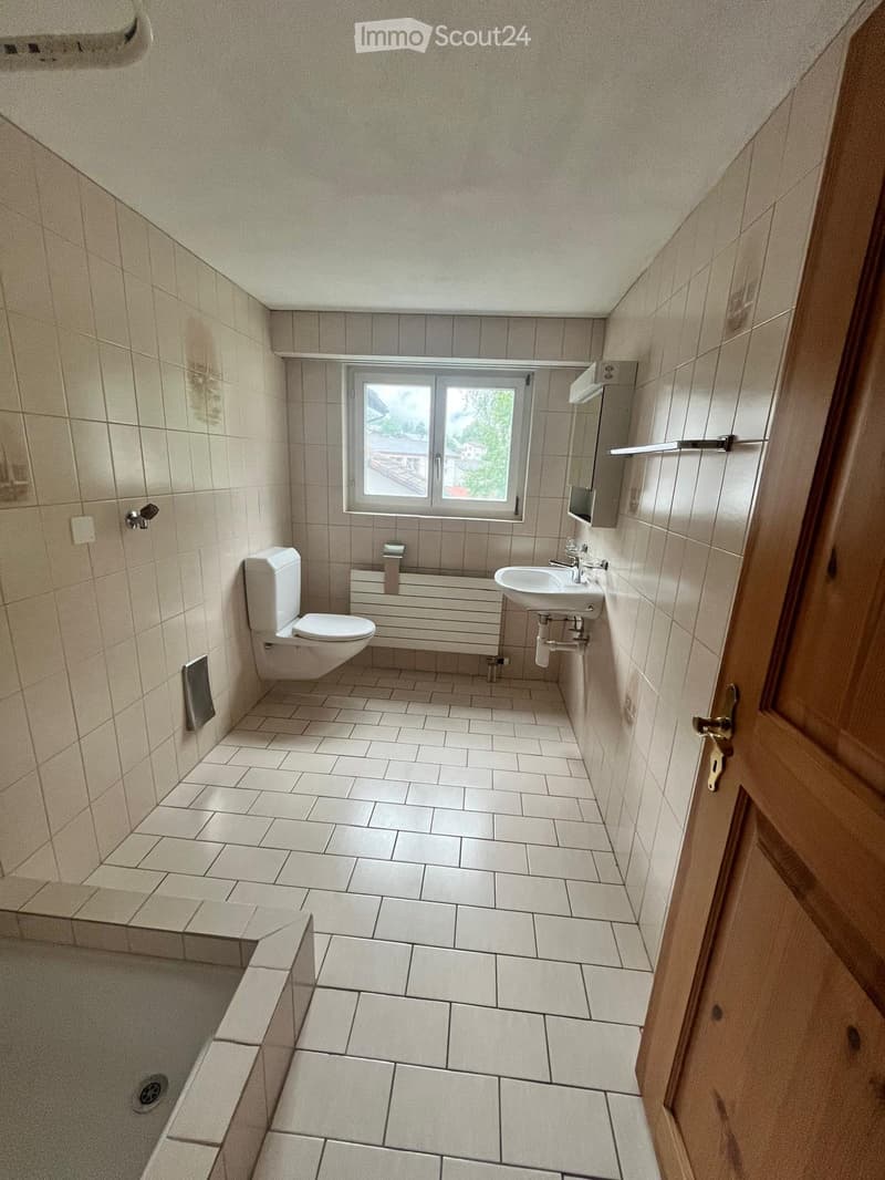 Bathroom with tiled walls and floor, toilet, sink, radiator, and a window offering a view of the outside.