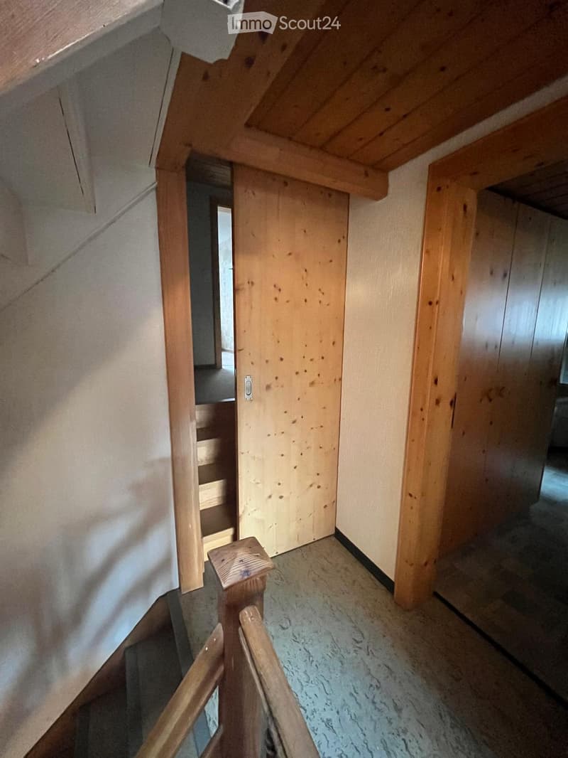 Wooden staircase with a handrail and open wooden door leading to another room. The walls are painted in a light color, and the floor is made of polished concrete.