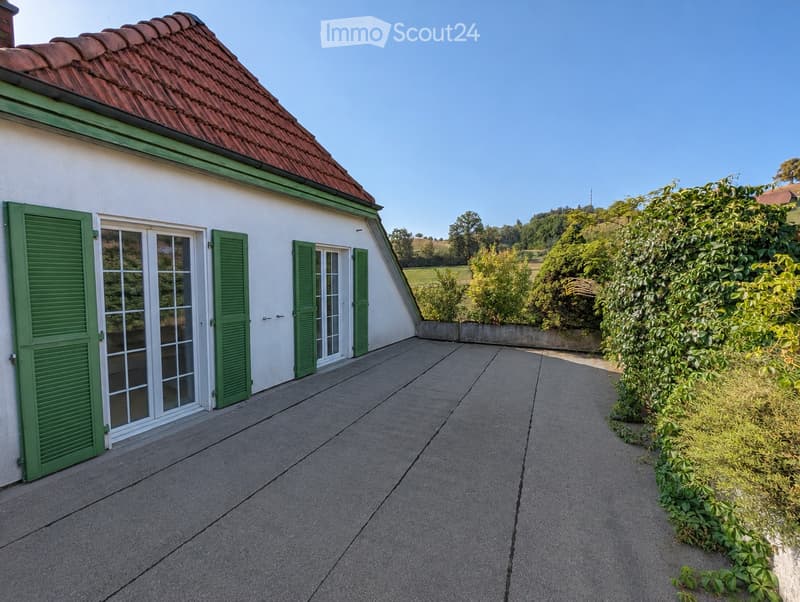 A single-story house with a red-tiled roof, white walls, and green shutters. The house features large windows and a concrete patio area with surrounding greenery.