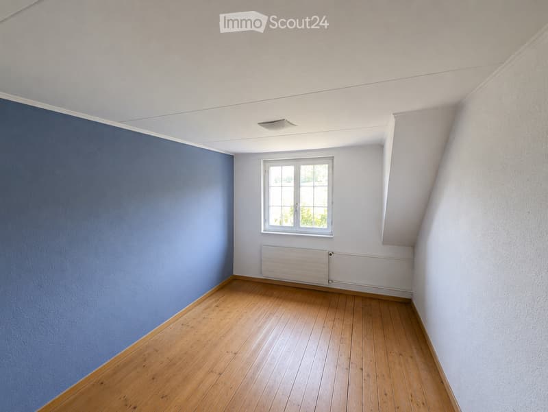 Empty room with wooden flooring, white walls, and a blue accent wall. Features a single window with white frame and a white radiator below it.