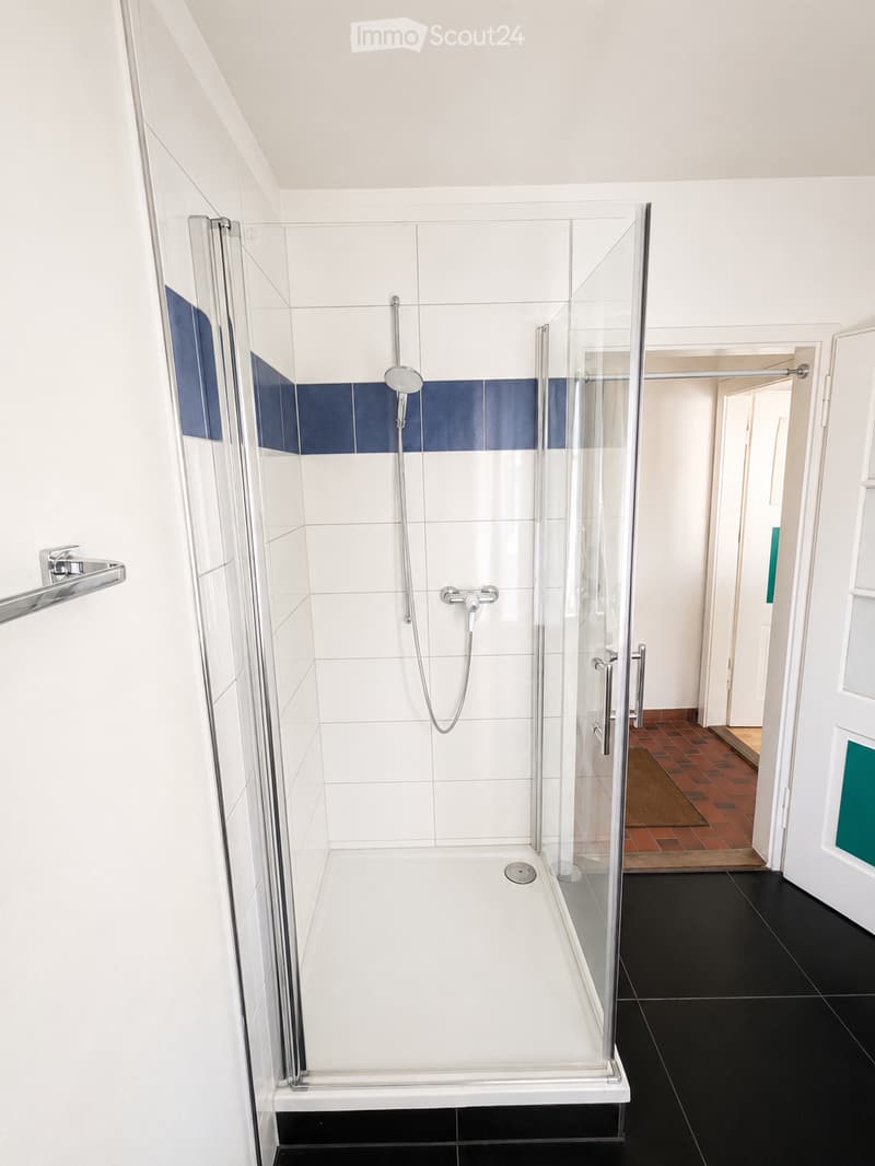 Modern bathroom with a glass-enclosed shower stall, white tiles, and blue accent tiles. Features a black tiled floor and a towel rack on the wall.