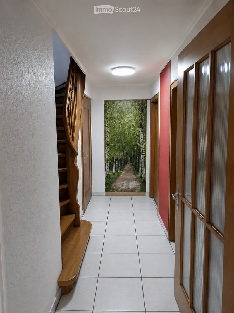 A clean, well-lit hallway with white tiled flooring, wooden doors, and a staircase leading upwards. The walls are painted in neutral tones, and a mural of a forest path is visible at the end of the corridor.