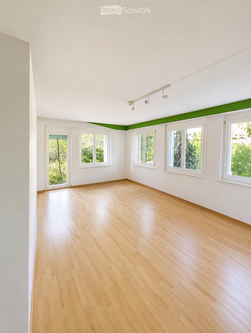 Empty room with light wooden flooring, white walls, and multiple windows offering natural light. The ceiling features a green accent strip and track lighting.