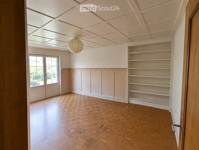 Empty room with wooden herringbone flooring, built-in wooden shelving unit, large windows, and a decorative ceiling light fixture.