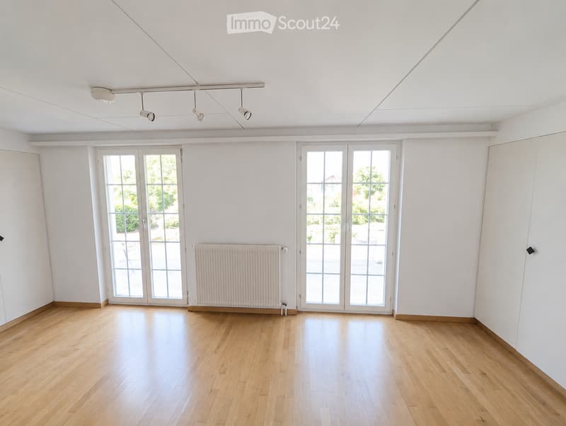 Empty room with light wooden flooring, white walls, large glass doors, and a radiator. No furniture or equipment present.