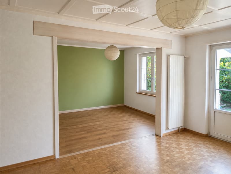 Empty room with wooden flooring, white walls, and large windows. Green accent wall visible in adjacent space. No furniture or equipment present.