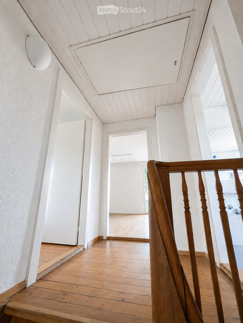 A bright, wooden-floored hallway with white walls and a wooden staircase railing. The hallway features a large, round wall-mounted mirror and leads to an open room with wooden flooring and white walls.