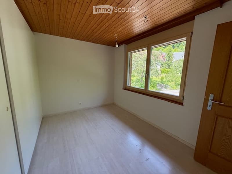 Empty room with light wooden flooring, white walls, and large windows with wooden frames. The ceiling has a wooden panel design.