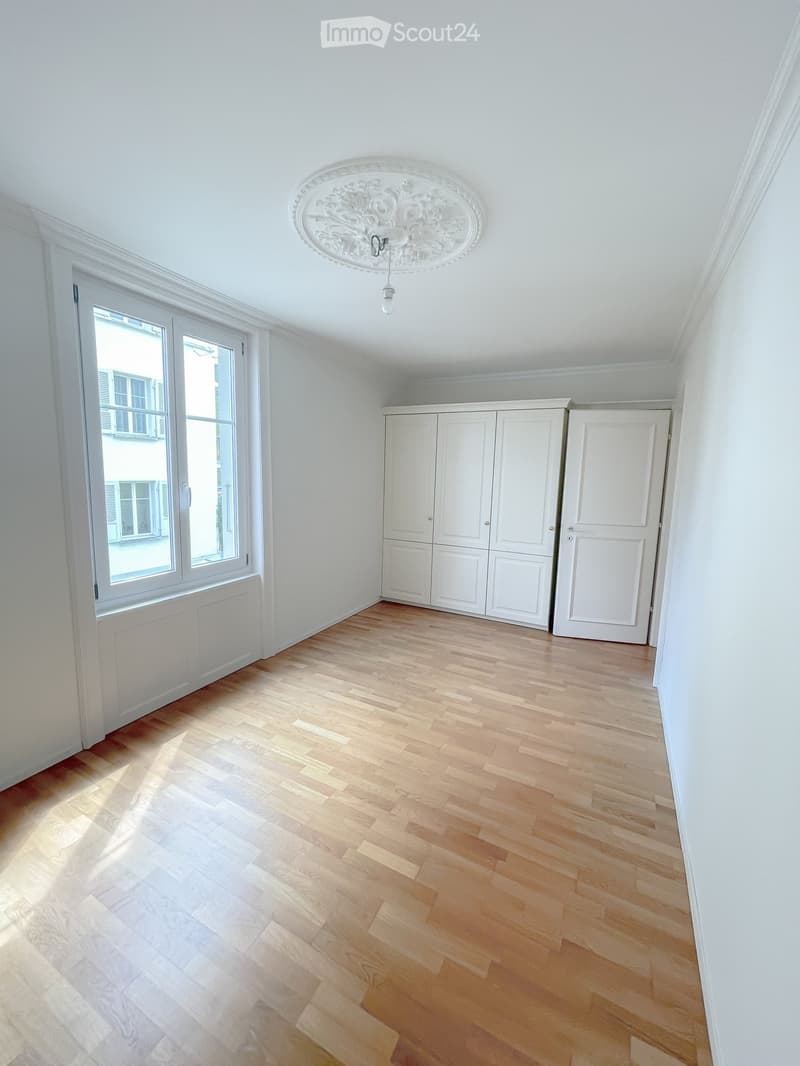 Empty room with light wooden flooring, white walls, large window, built-in white cabinetry, and a decorative ceiling light fixture.