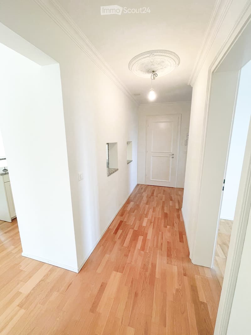 Empty hallway with light wooden flooring, white walls, and a single closed door at the end. A decorative ceiling light fixture is visible, and there are small mirrors mounted on the walls.