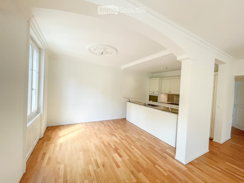 Bright, open-plan living and dining area with a modern kitchen featuring white cabinets, a granite countertop, and a built-in oven. The space has wooden flooring and large windows allowing natural light.
