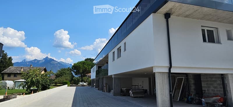 Modern two-story building with white facade, large windows, and a raised ground floor. The background features a scenic mountain view.