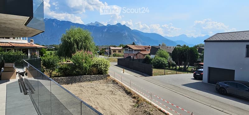 View from a modern balcony overlooking a residential area with houses, trees, and distant mountains.