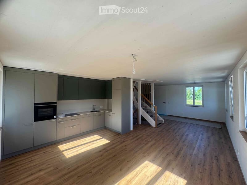 Empty room with wooden flooring, white walls, and modern kitchen cabinets visible in the background. Natural light streams in from a window, and a staircase leads to another level.