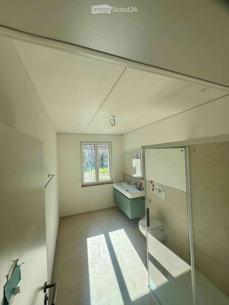 Modern bathroom with a sink, mirror, toilet, and a glass-enclosed shower stall. The room features light-colored tiles and a window allowing natural light.