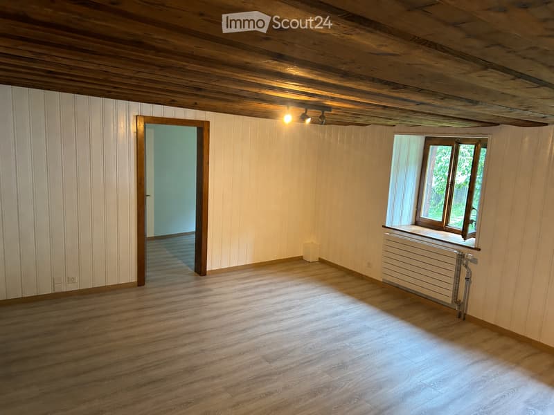 Empty room with wooden flooring, white paneled walls, exposed wooden ceiling beams, and a single window with a radiator below it. A small white box is placed near the window.