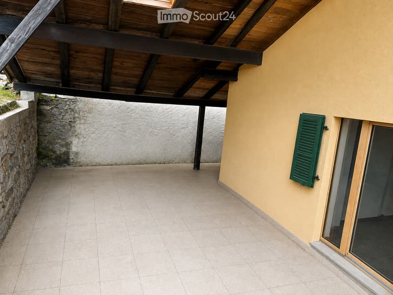 Covered outdoor area with tiled floor, wooden ceiling beams, stone wall, and green shutter on the adjacent building.