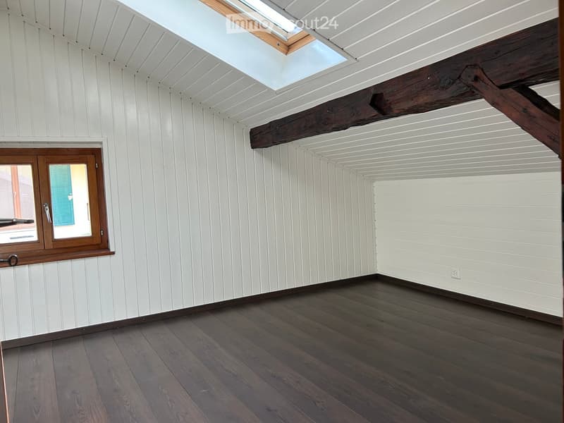 Empty room with white paneled walls, dark wooden floor, exposed wooden beams, and a skylight window.