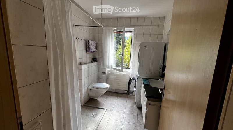 Bathroom with a toilet, washing machine, sink, mirror, radiator, and a window with a curtain. A towel is hanging on the wall.
