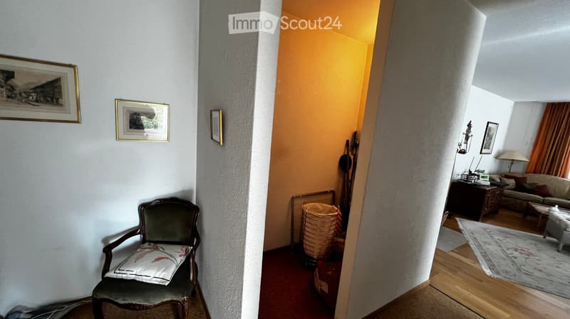 A furnished room with a wooden chair, sofa, coffee table, and a table lamp. The room features a mix of wooden and carpeted flooring, with framed pictures on the walls.