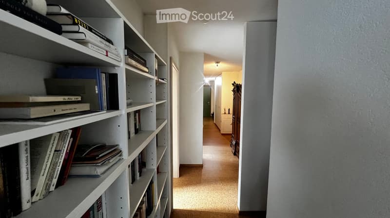 A narrow hallway with light-colored walls and a wooden floor, leading to a room with a bookshelf filled with books and a small cabinet.