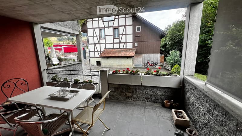 Balcony with a table, chairs, and flower boxes, overlooking a traditional half-timbered house and greenery.