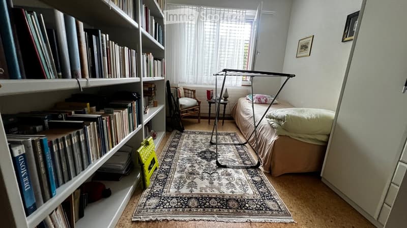 Cozy bedroom with a bed, bookshelf filled with books, a small table, a chair, and a patterned rug on the floor.