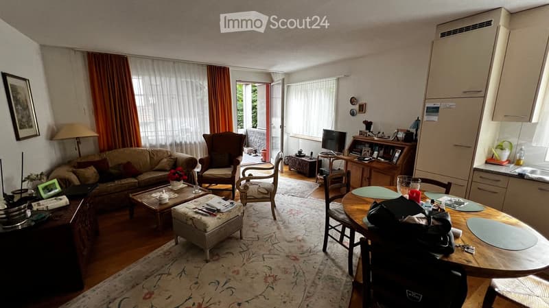 A well-furnished living and dining area with a sofa, coffee table, ottoman, chairs, dining table, and kitchen cabinetry. The space features a patterned rug, wooden flooring, and large windows with curtains.