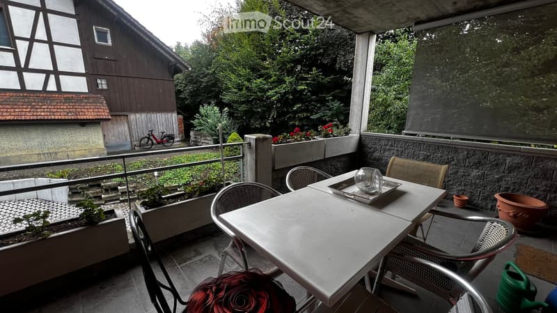 Balcony with a table, chairs, and potted plants, overlooking a garden area with a traditional house in the background.