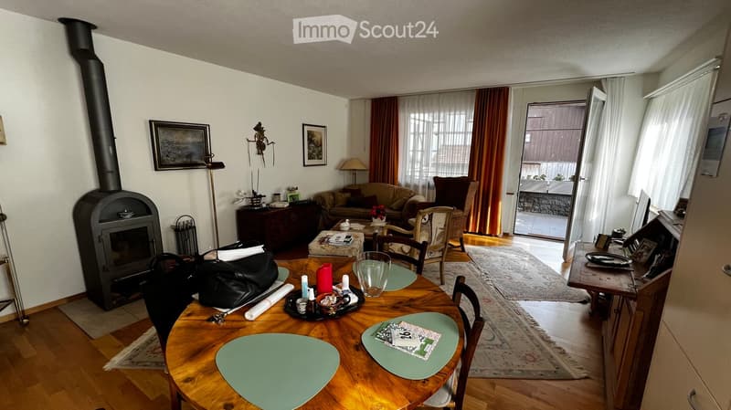 A cozy living room with a fireplace, a sofa, a wooden dining table, chairs, and a lamp. The room features large windows with curtains and a view of the balcony.
